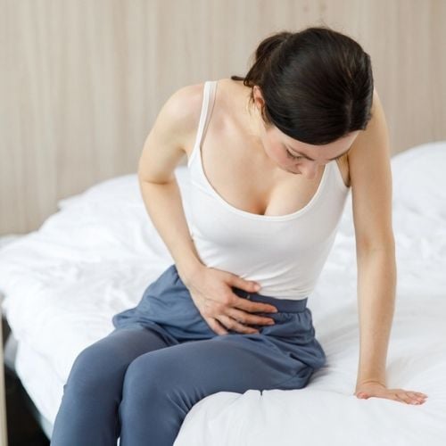 Woman sitting on bed, experiencing abdominal pain, relevant to vaginal mesh complications and symptoms like chronic pelvic pain.